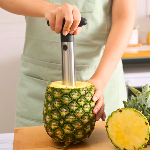 stainless steel and black handheld pineapple coring/slicing tool on a whole pineapple on a wooden cutting board, with a cut pineapple half and green leaves visible.